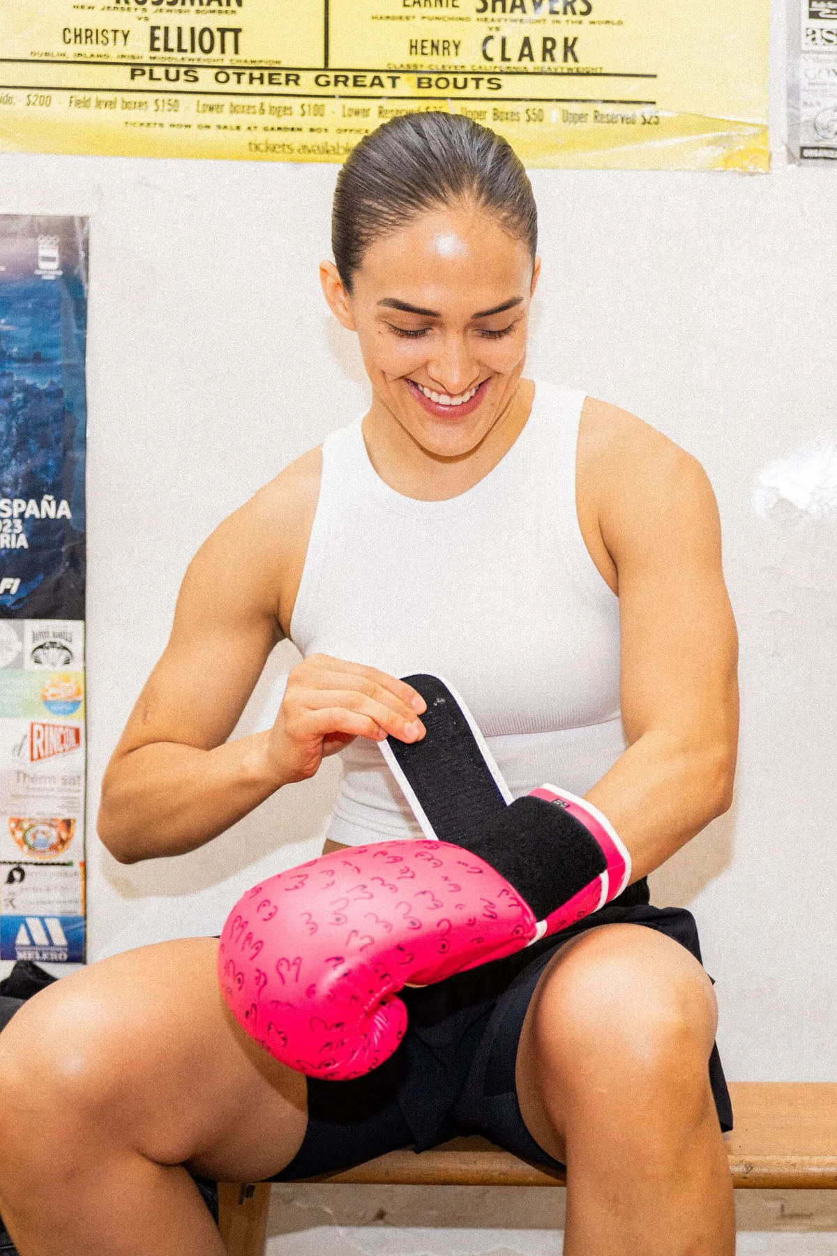Boxeadora sonriendo mientras se cierra el guante rosa en la muñeca, sentada en un banco del gimnasio