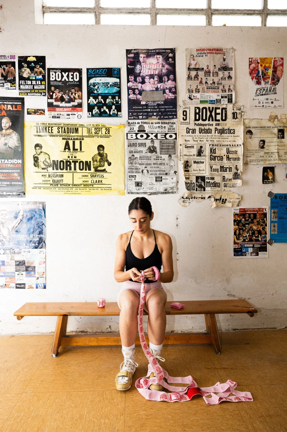 Boxeadora vendándose las manos en un banco del gimnasio, bajo carteles de boxeo